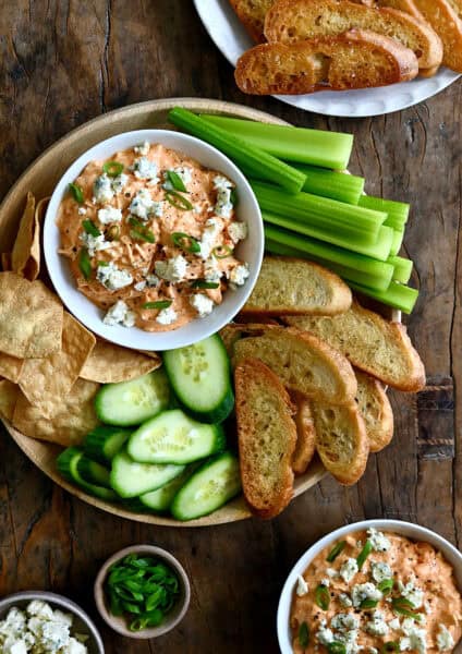 Buffalo chicken dip in a bowl on a plate surrounded by baguette toasts, sliced cucumbers, tortilla chips and celery sticks.