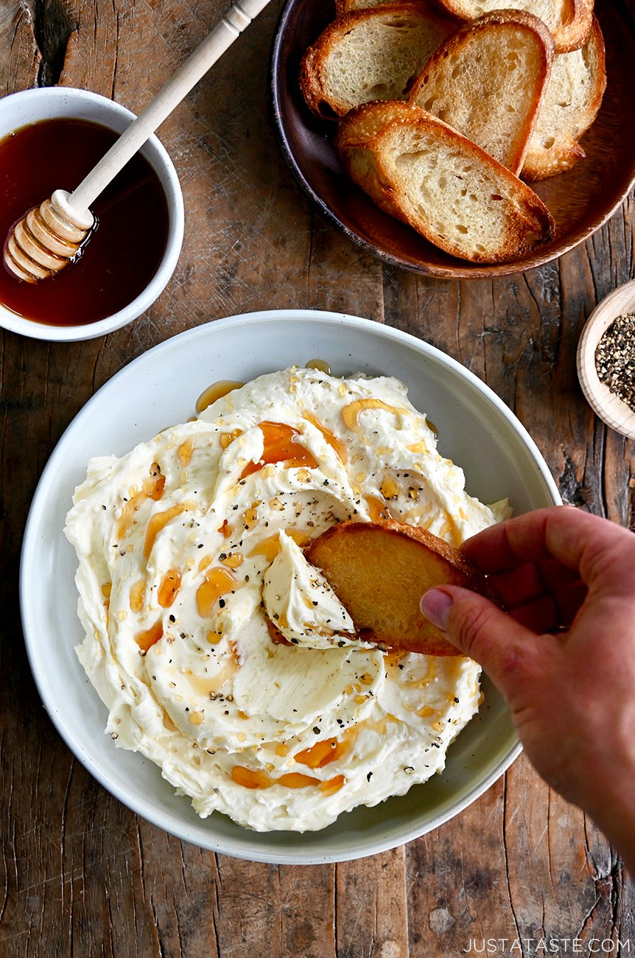 A hand dips a toasty baguette into whipped Brie that's been garnished with cracked black pepper and drizzled with honey. 