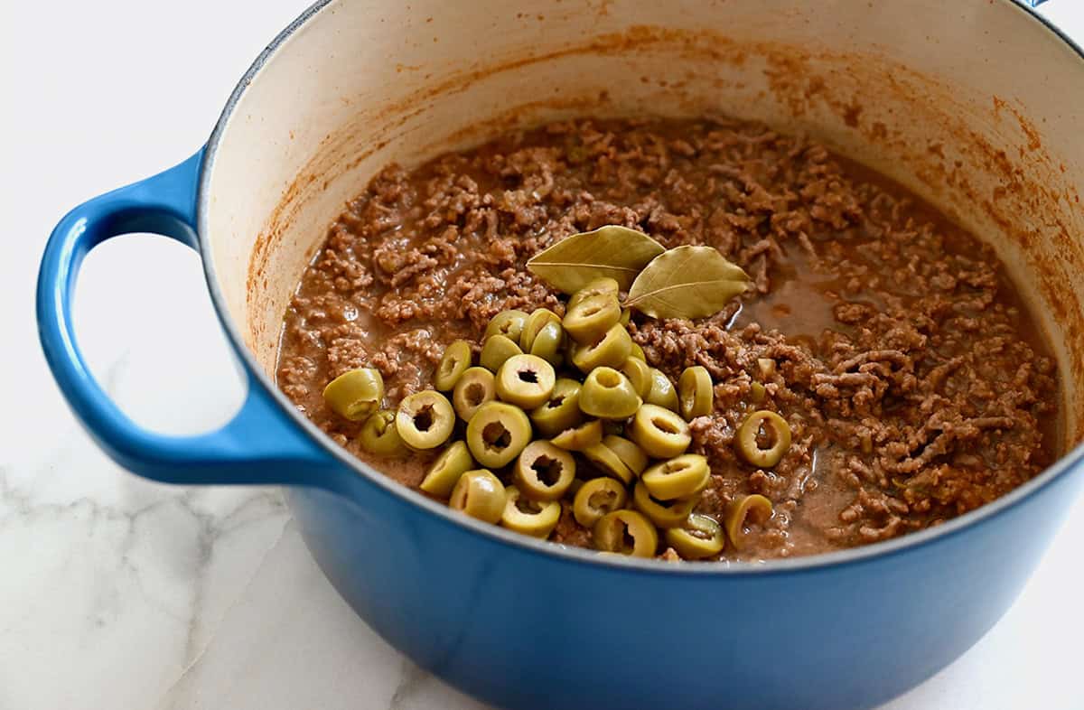 A large stockpot containing ground beef, green olives and two bay leaves.