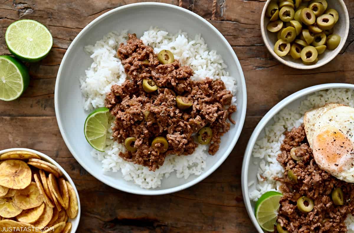 Cuban picadillo (ground beef) with green olives and raisins atop steamed white rice in a bowl next to a small dish containing plantain chips.