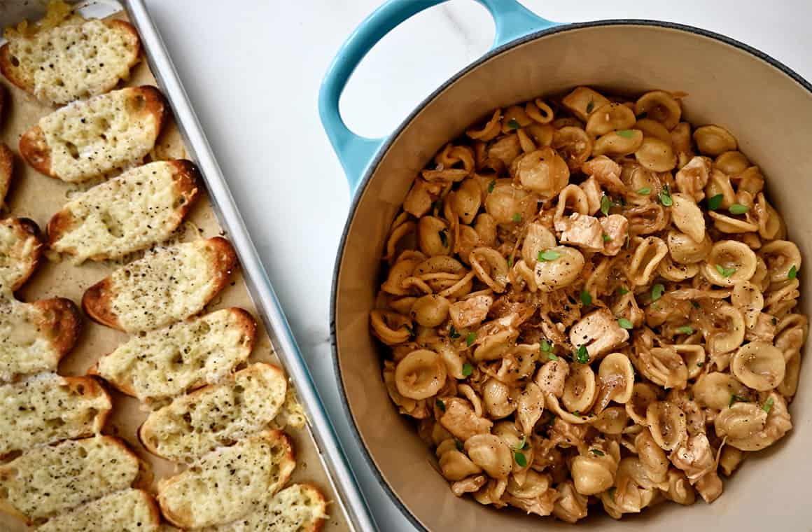 A top-down view of French onion chicken pasta in a stockpot next to a baking sheet with cheesy baguette toasts
