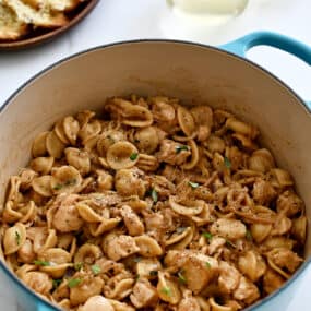 A blue stockpot containing French onion orecchiette pasta with chicken next to a plate topped with cheesy baguette toasts