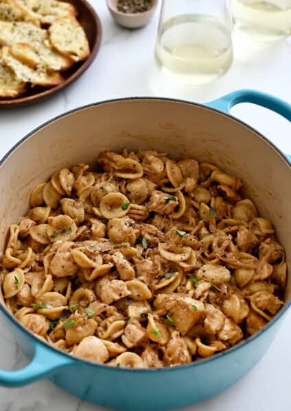 A blue stockpot containing French onion orecchiette pasta with chicken next to a plate topped with cheesy baguette toasts