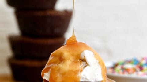 A fudgy brownie cupcake topped with a scoop of vanilla ice cream being drizzled with caramel sauce