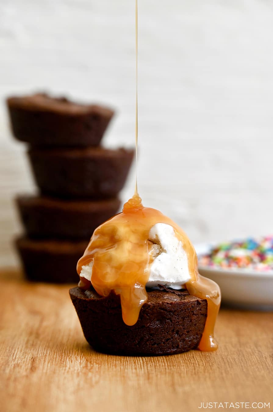 A fudgy brownie cupcake topped with a scoop of vanilla ice cream being drizzled with caramel sauce