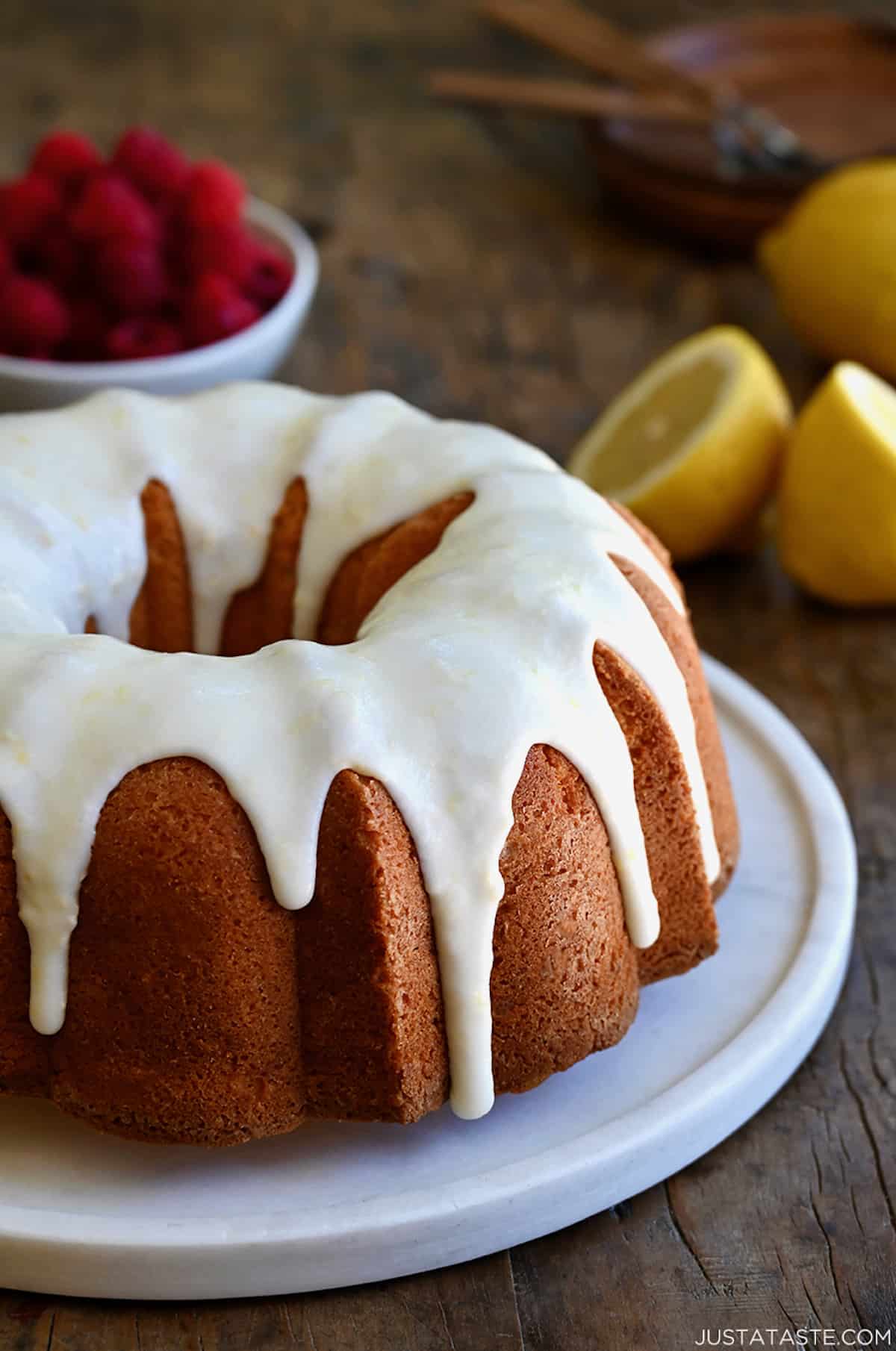 A glazed lemon pound cake on a round, white serving platter with a bowl filled with raspberries in the background.