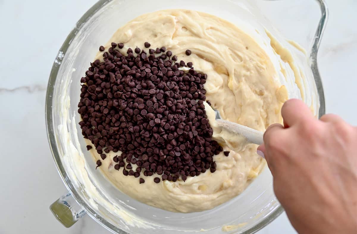A hand holding a spatula mixes mini chocolate chips into cake batter in a clear bowl.