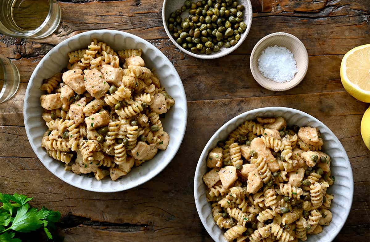 Two bowls containing chicken piccata pasta with capers next to a small bowl filled with sea salt and a lemon cut in half.