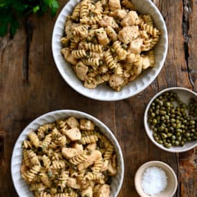 A top-down view of two bowls containing Chicken Piccata Pasta next to two small bowls containing capers and sea salt.