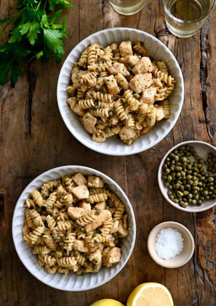 A top-down view of two bowls containing Chicken Piccata Pasta next to two small bowls containing capers and sea salt.