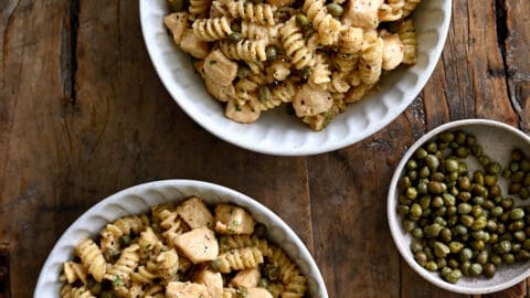 A top-down view of two bowls containing Chicken Piccata Pasta next to two small bowls containing capers and sea salt.