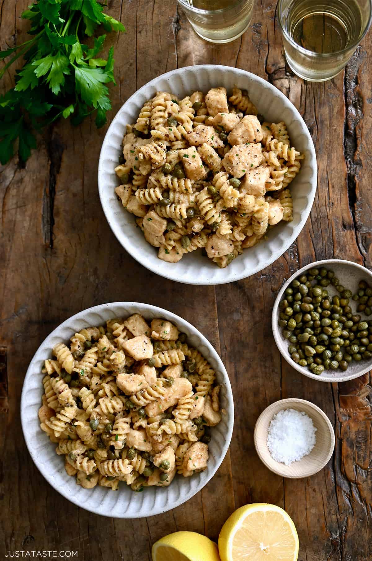 A top-down view of two bowls containing Chicken Piccata Pasta next to two small bowls containing capers and sea salt.