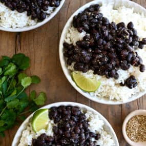 Three plates containing white rice topped with Cuban black beans and a lime wedge next to a bunch of cilantro.
