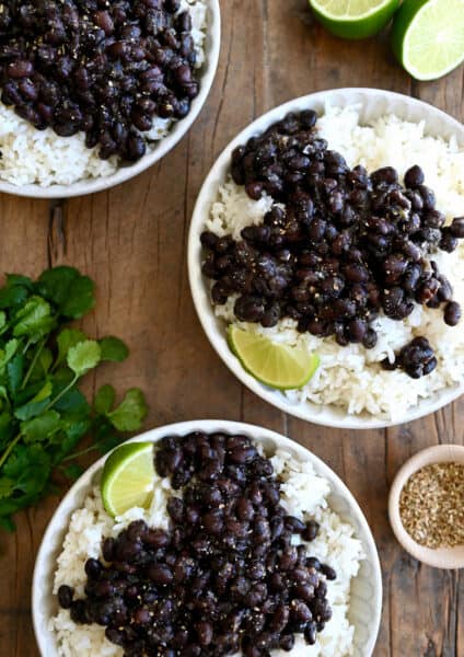 Three plates containing white rice topped with Cuban black beans and a lime wedge next to a bunch of cilantro.