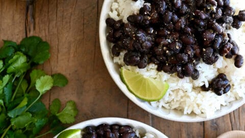 Three plates containing white rice topped with Cuban black beans and a lime wedge next to a bunch of cilantro.