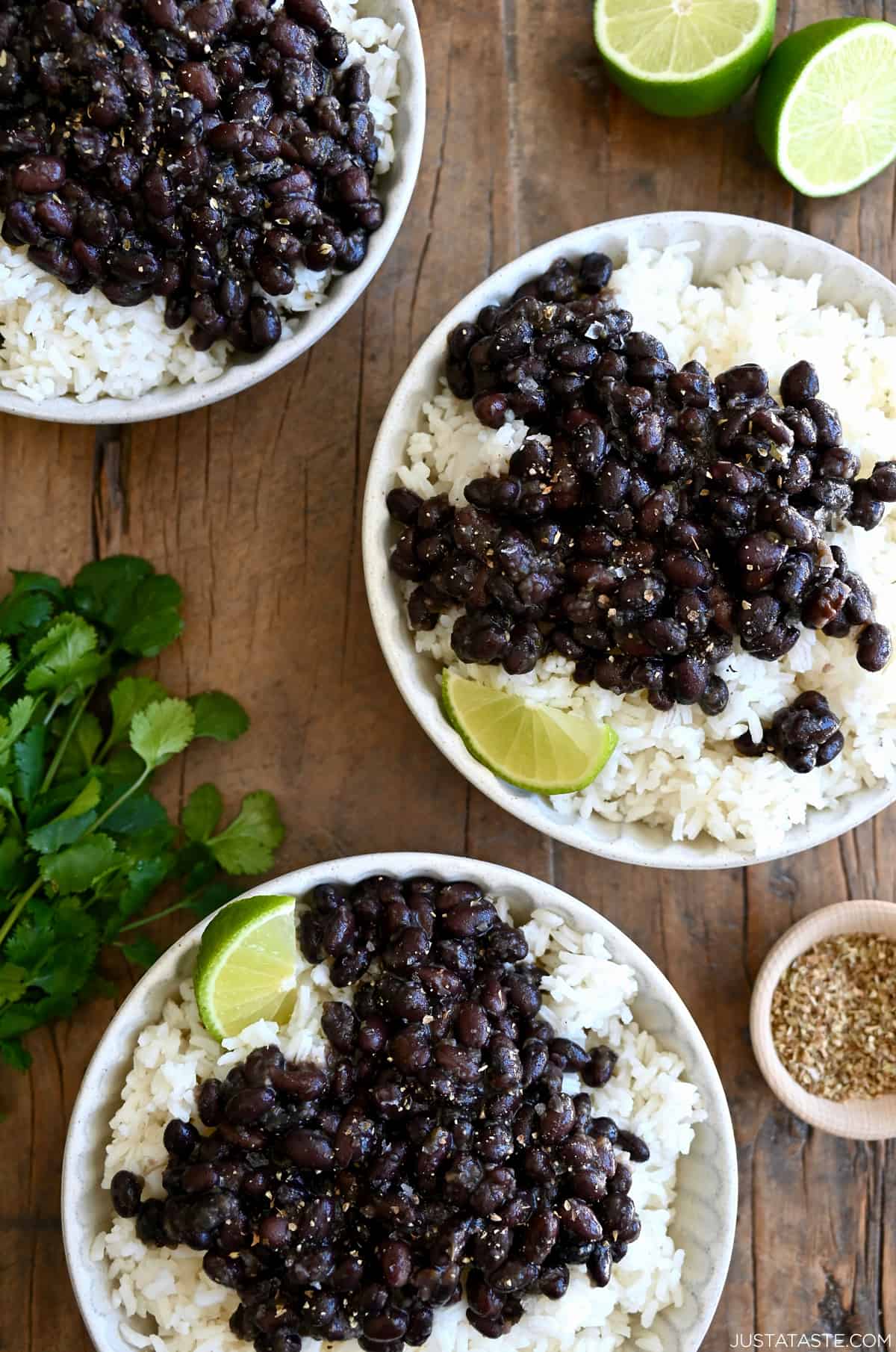 Three plates containing white rice topped with Cuban black beans and a lime wedge next to a bunch of cilantro.