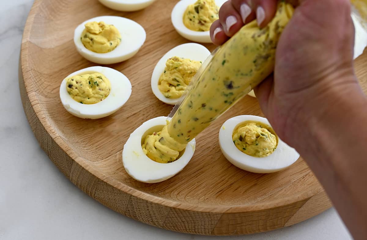 Deviled egg filling being piped into halved eggs on a wood tray.