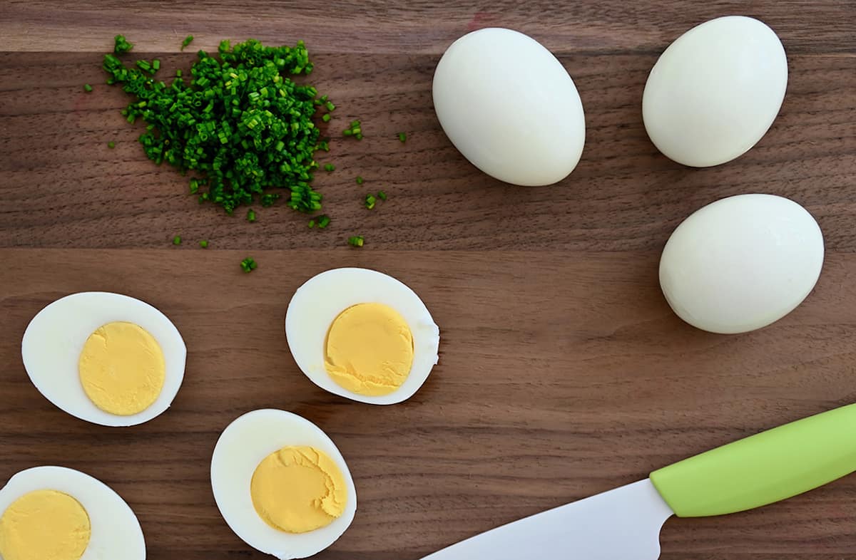 Hard boiled eggs on a cutting board with chopped fresh chives and a sharp knife.