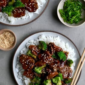 Quick Caramelized Pork and Broccoli over a bed of rice on a plate with chopsticks next to a small bowl containing sesame seeds and a bowl containing broccoli florets.