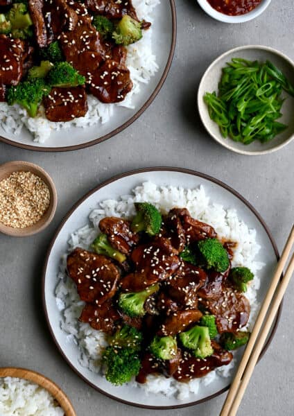Quick Caramelized Pork and Broccoli over a bed of rice on a plate with chopsticks next to a small bowl containing sesame seeds and a bowl containing broccoli florets.