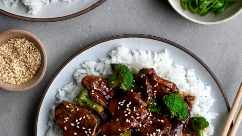 Quick Caramelized Pork and Broccoli over a bed of rice on a plate with chopsticks next to a small bowl containing sesame seeds and a bowl containing broccoli florets.