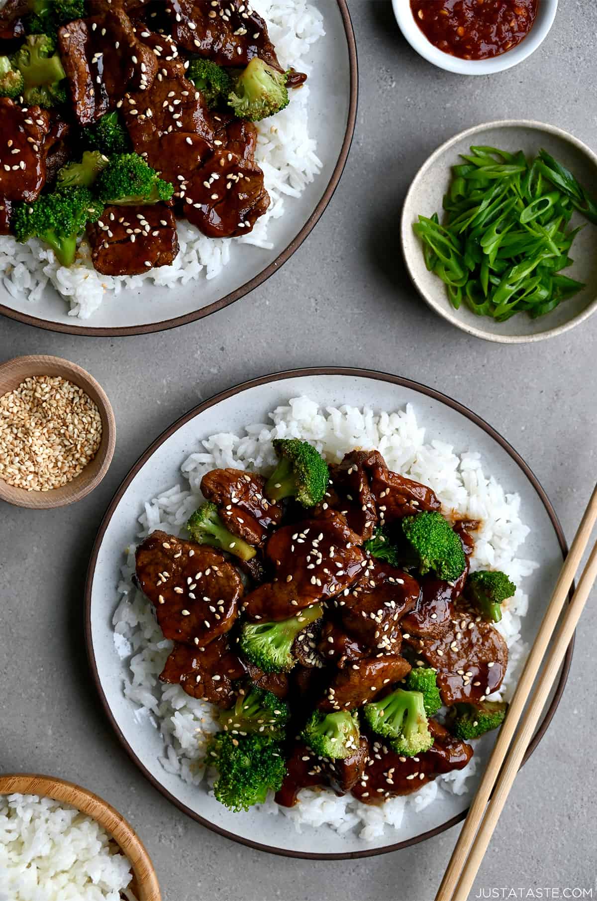 Quick Caramelized Pork and Broccoli over a bed of rice on a plate with chopsticks next to a small bowl containing sesame seeds and a bowl containing broccoli florets.