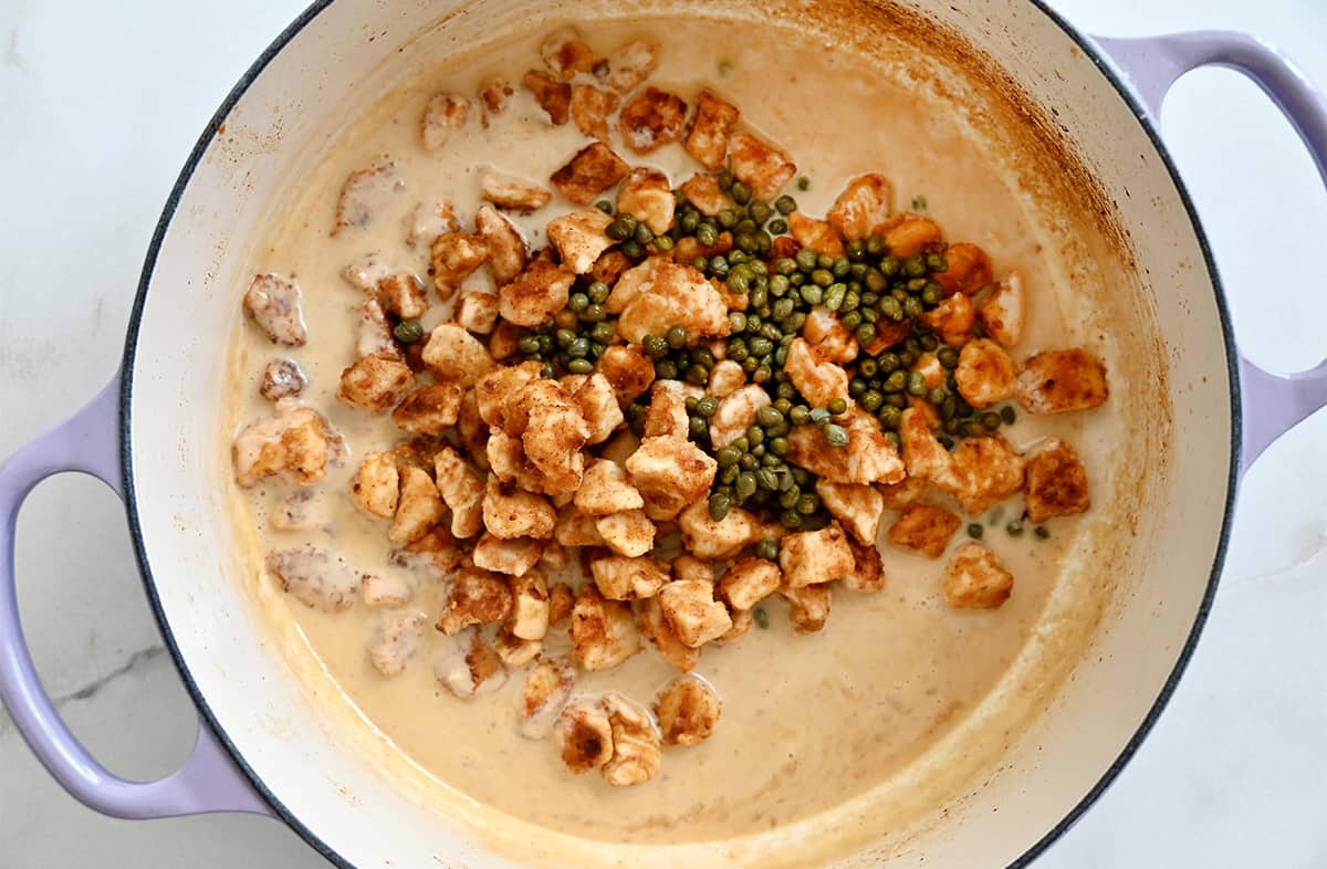 A top-down view of a large saucepan with a creamy sauce, chicken pieces and capers.