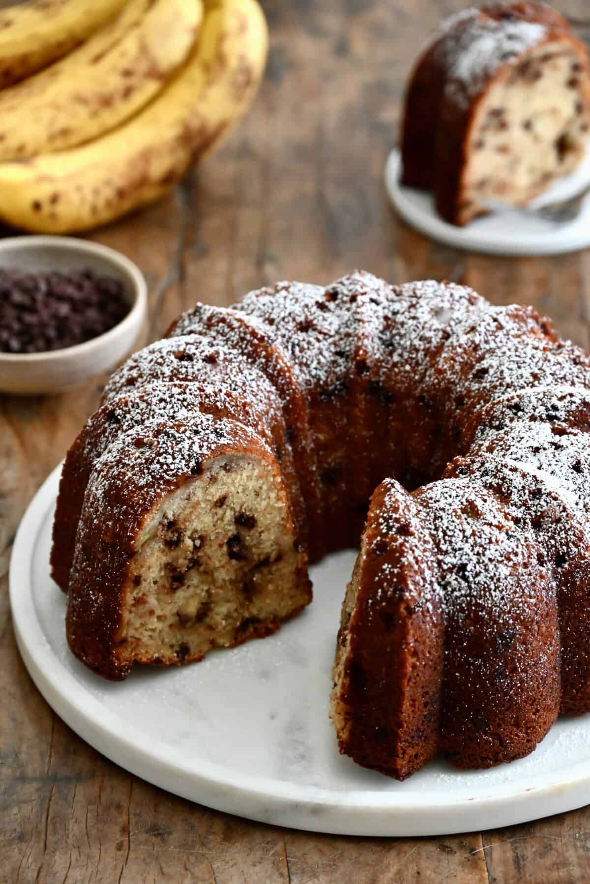 A banana chocolate chip bundt cake with one slice missing, dusted with powdered sugar.