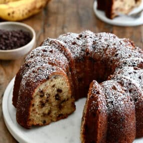 A banana chocolate chip bundt cake with one slice missing, dusted with powdered sugar.