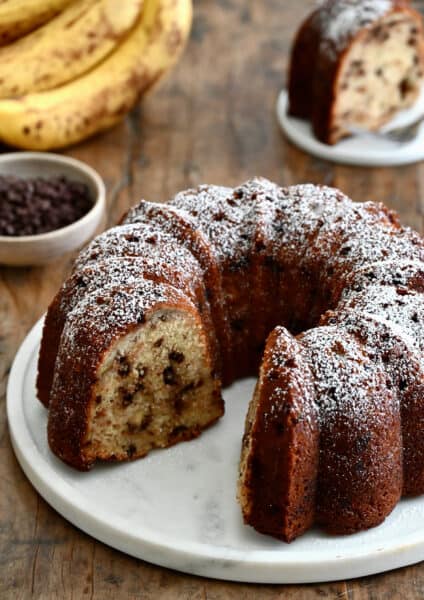 A banana chocolate chip bundt cake with one slice missing, dusted with powdered sugar.