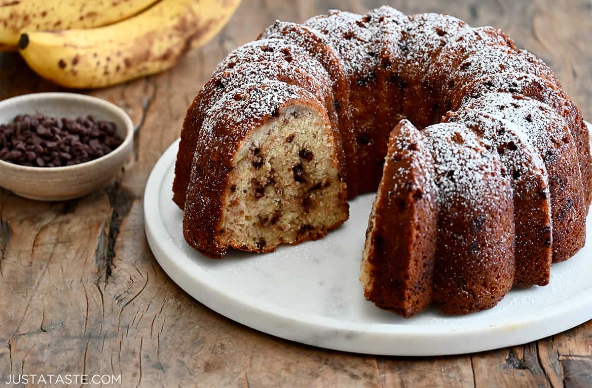 A banana bundt cake studded with mini chocolate chips with a slice missing on a marble cake platter.