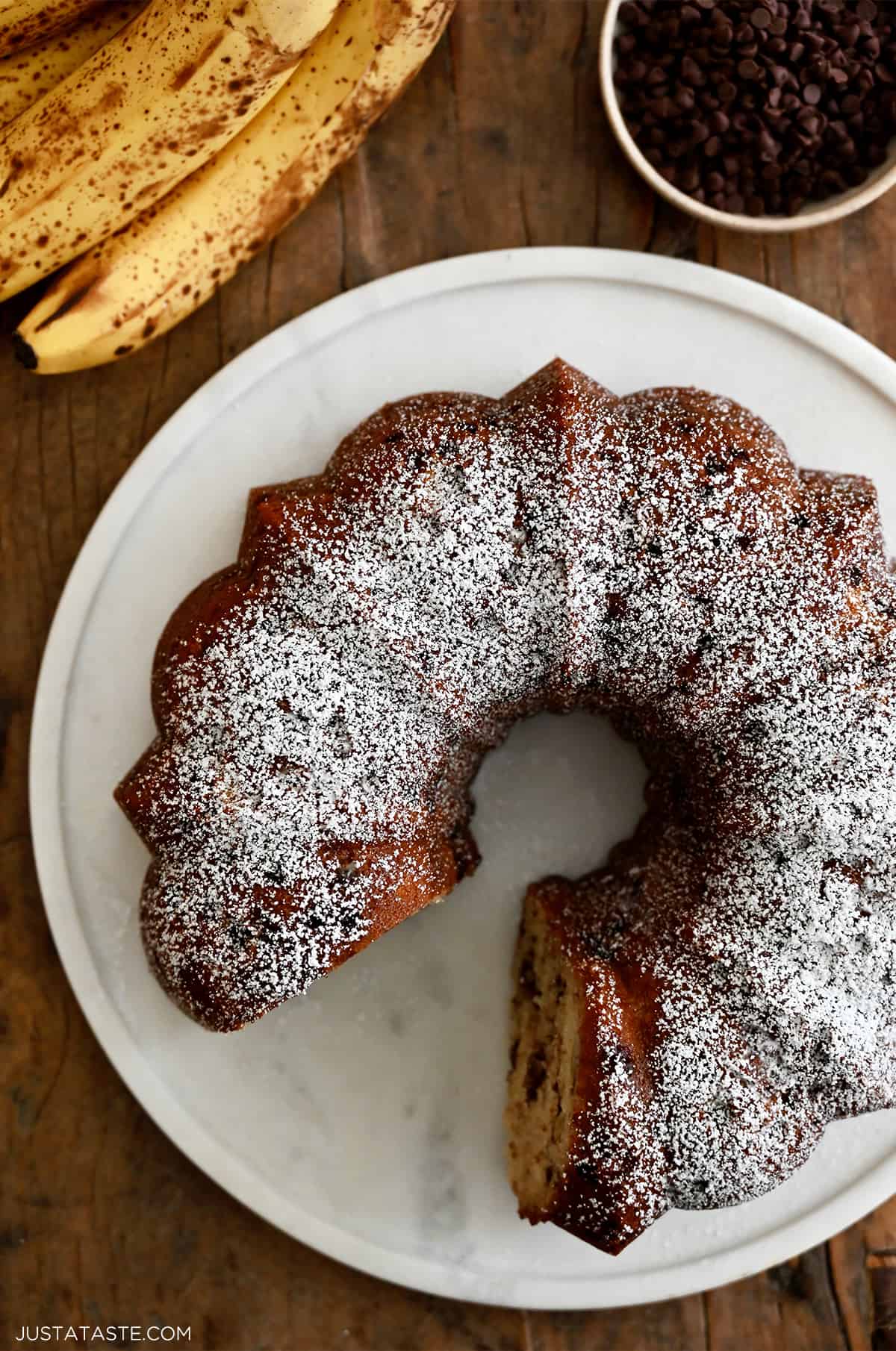 A top-down view of a chocolate chip banana bundt cake dusted with powdered sugar on a round cake platter next to a bunch of overripe bananas and a bowl containing mini chocolate chips.