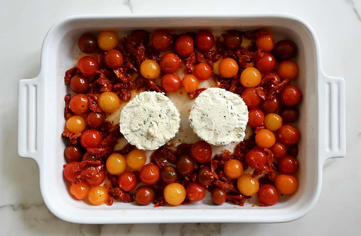 A top-down view of a white baking dish with two Boursin cheese rounds surrounded by cherry tomatoes and sun-dried tomatoes.