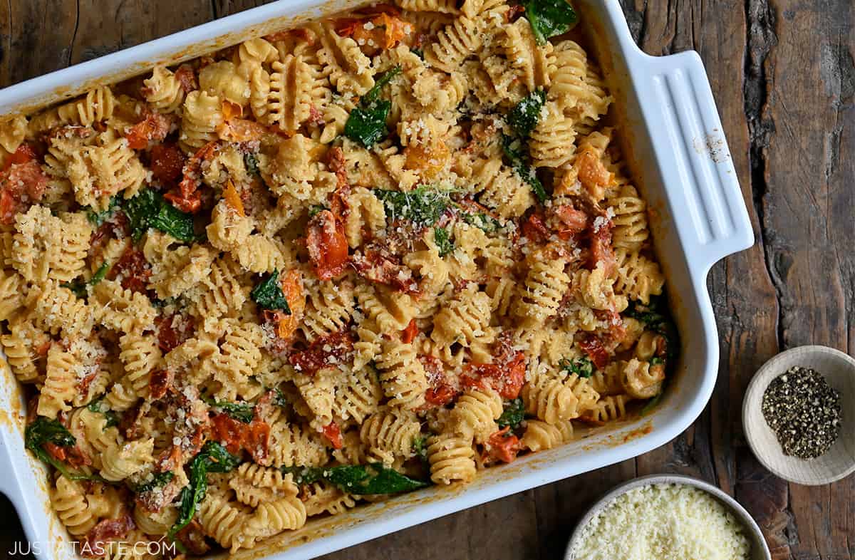 A top-down view of Baked Boursin Pasta with roasted tomatoes and spinach in a white baking dish next to small bowls containing black pepper and Parmesan cheese.