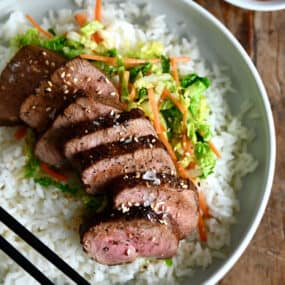 Sliced pork tenderloin over veggies and a bed of rice on a dinner plate with chopsticks.