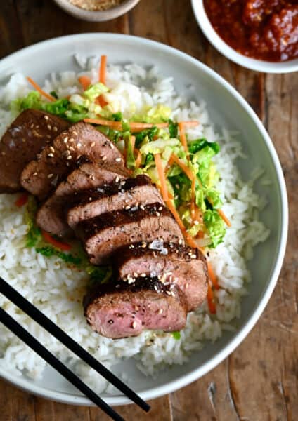 Sliced pork tenderloin over veggies and a bed of rice on a dinner plate with chopsticks.