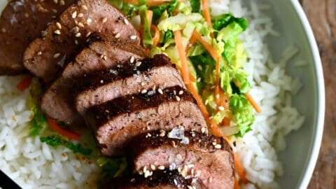 Sliced pork tenderloin over veggies and a bed of rice on a dinner plate with chopsticks.