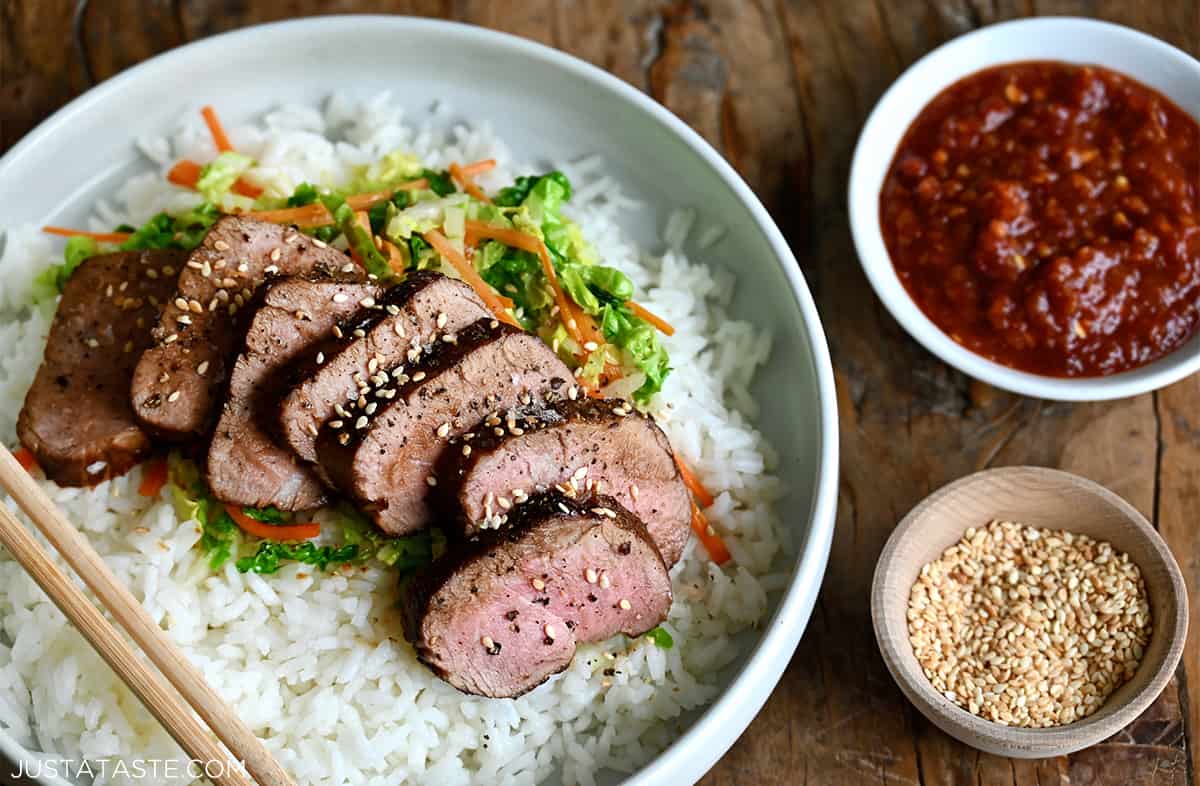 Sliced pork tenderloin over veggies and white rice on a plate next to small bowls containing sesame seeds and chili sauce.