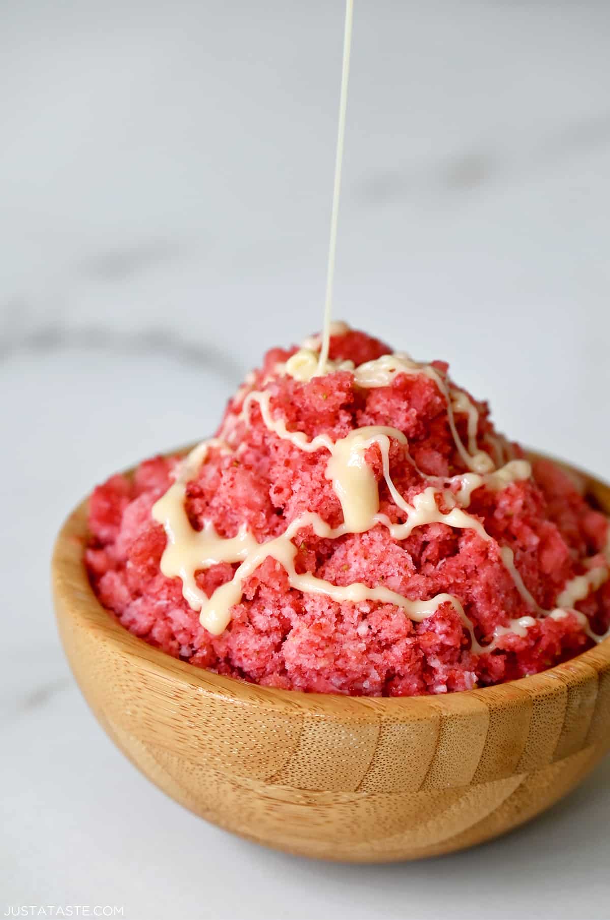 A wood bowl containing strawberry shaved ice with a drizzle of sweetened condensed milk