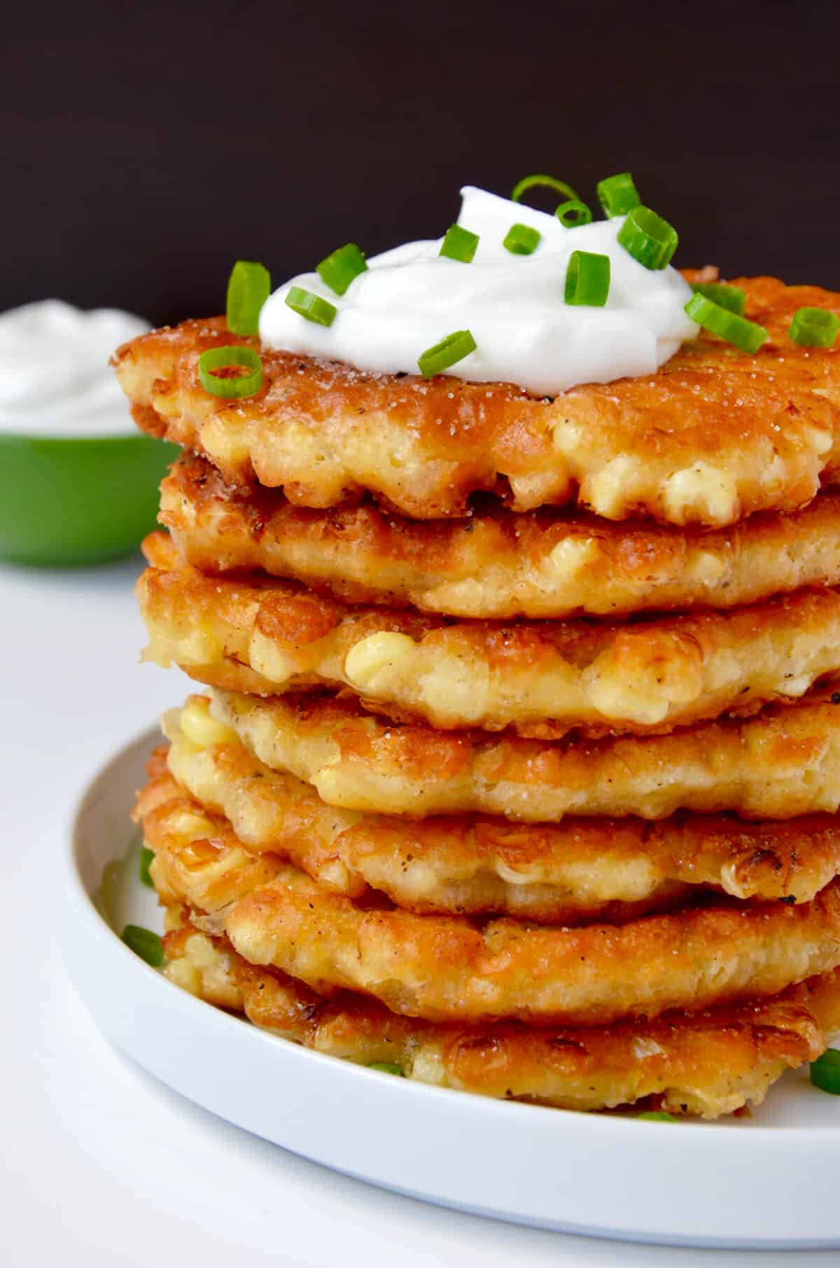 A tall stack of corn fritters topped with a dollop of sour cream and garnished with chopped scallions.