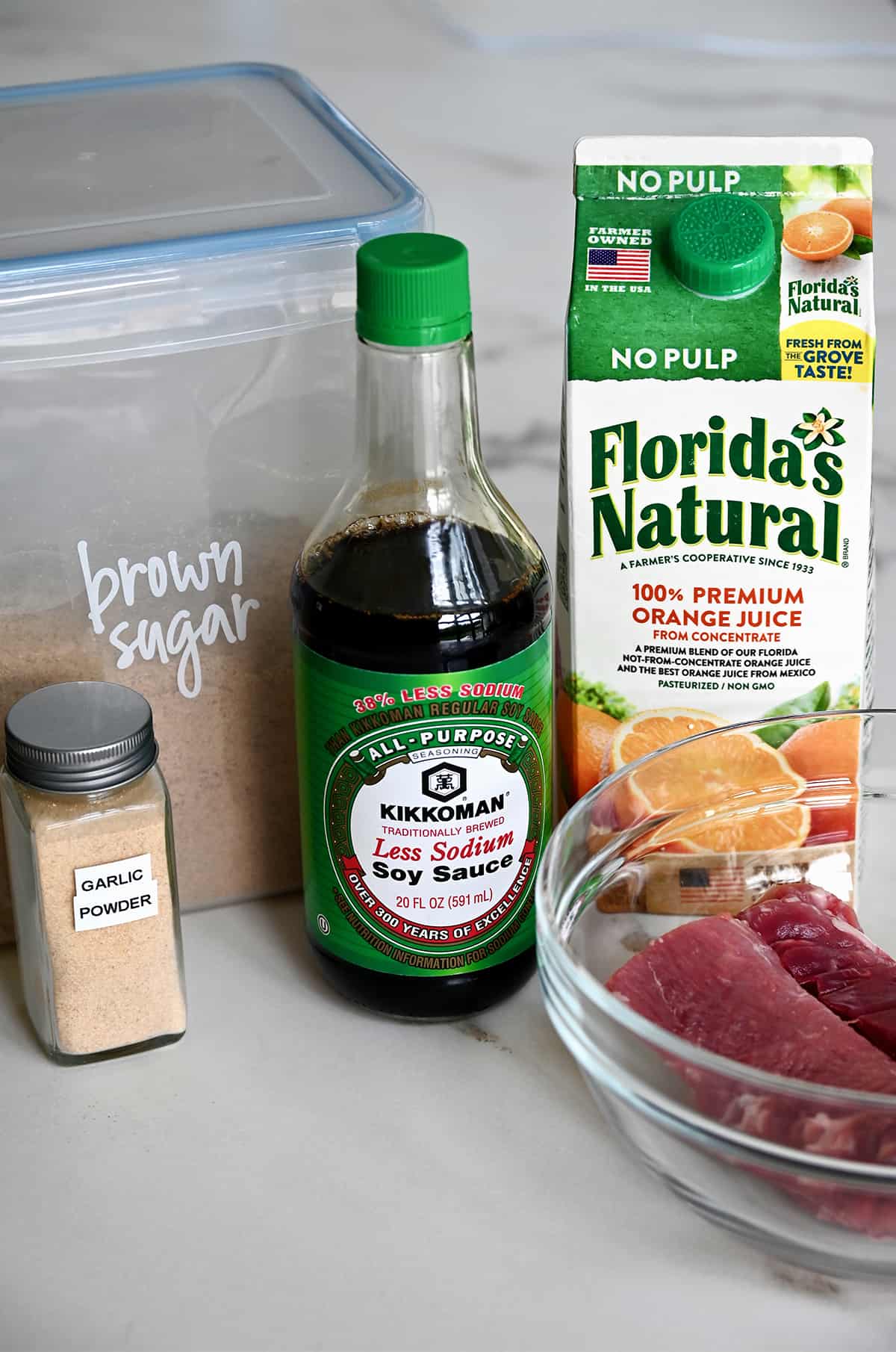 A small jar containing garlic powder next to a plastic container with brown sugar, a bottle of low sodium soy sauce, a carton of Florida's Natural orange juice and a clear bowl with raw pork tenderloin.