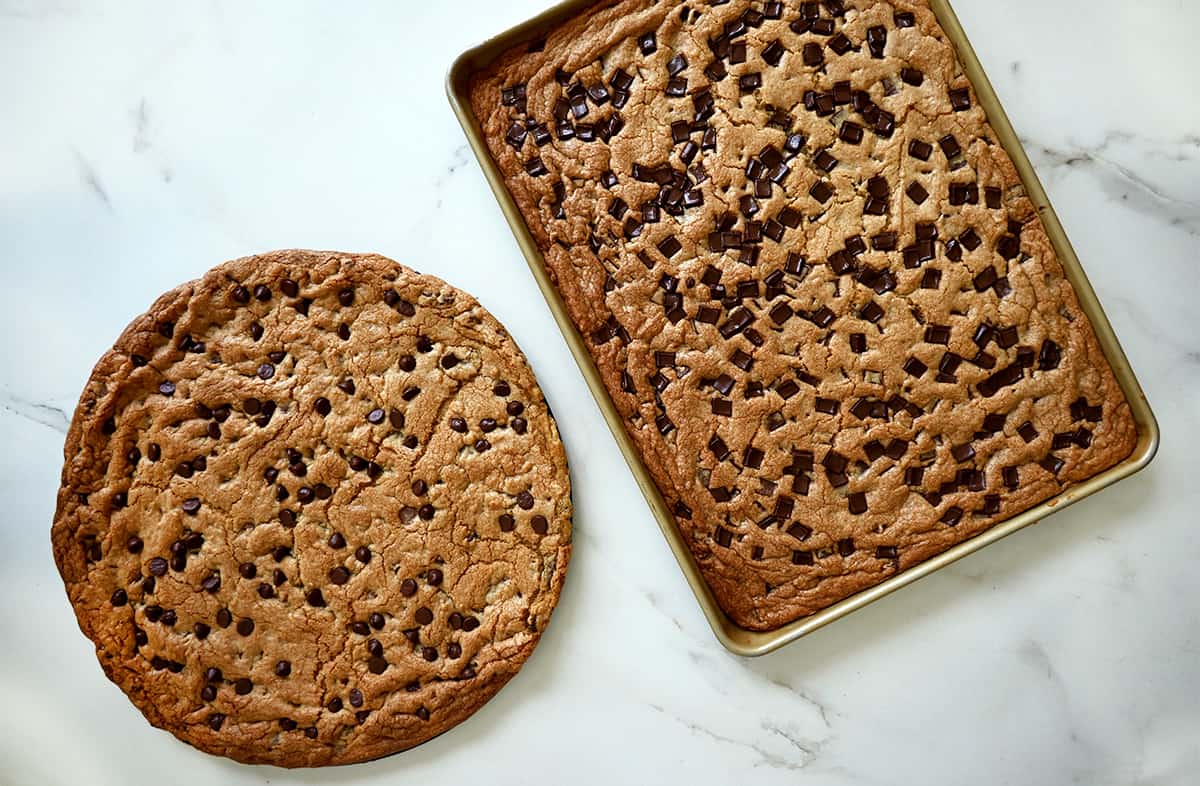 A top-down view of two baked cookie cakes, one on a round pan and the other on a baking sheet.