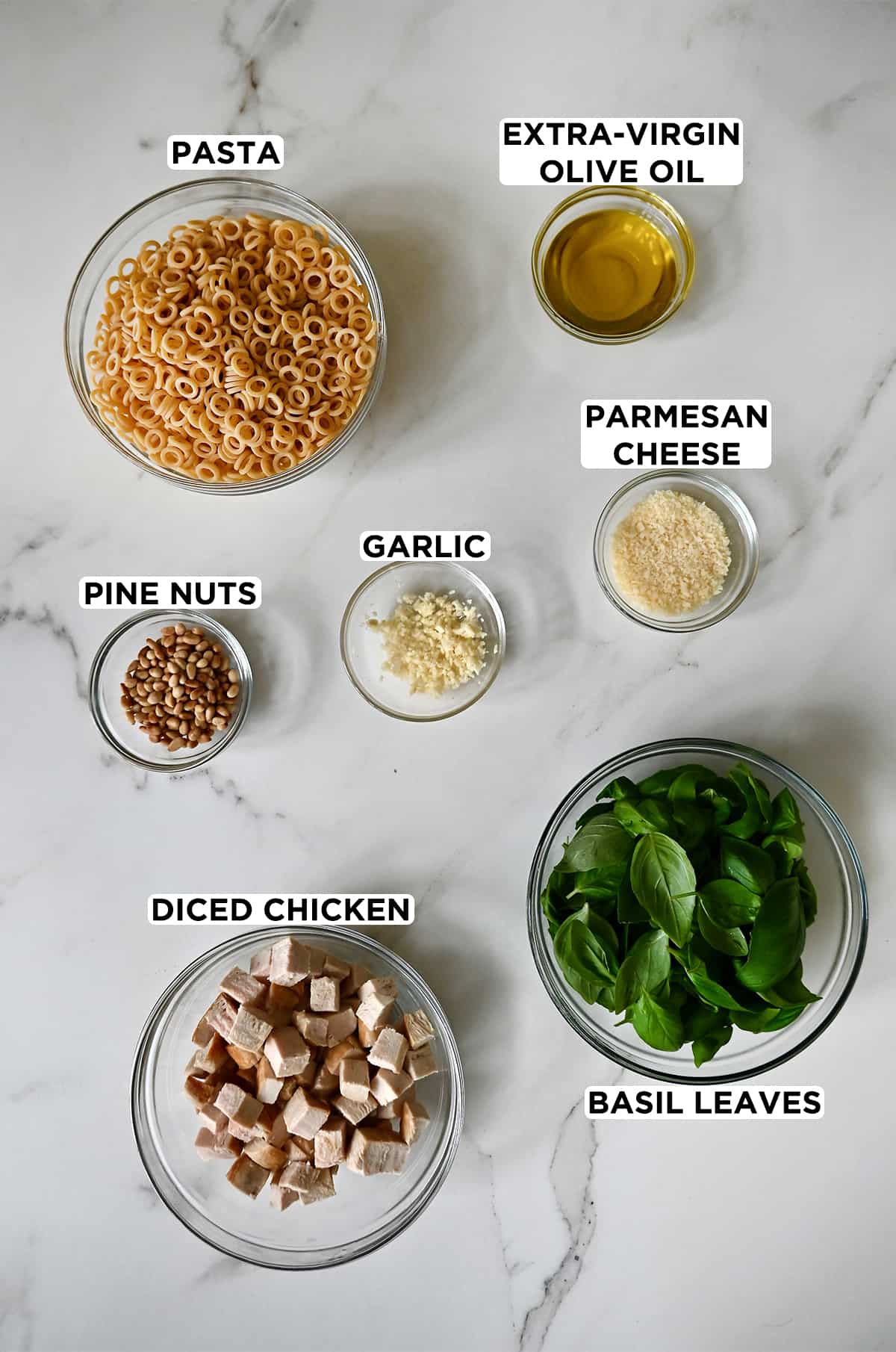 A top-down view of various sized clear bowls containing dry pasta, olive oil, parmesan cheese, basil, cubed cooked chicken breast, garlic and pine nuts.