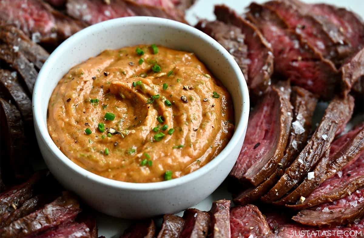 A small white bowl containing cowboy dipping sauce surrounded by sliced steak that's garnished with large-flake sea salt.
