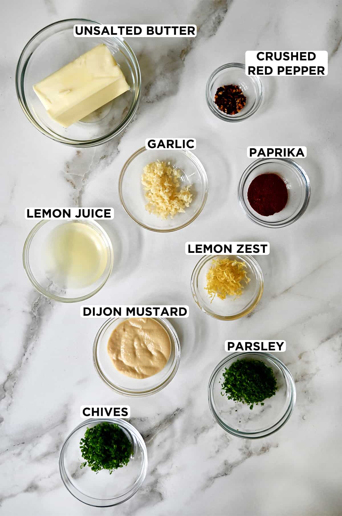 A top-down view of small glass bowls containing all of the ingredients to make cowboy butter, including butter, minced garlic, crushed red pepper flakes, paprika, lemon juice and zest, dijon mustard, parsley and chives.