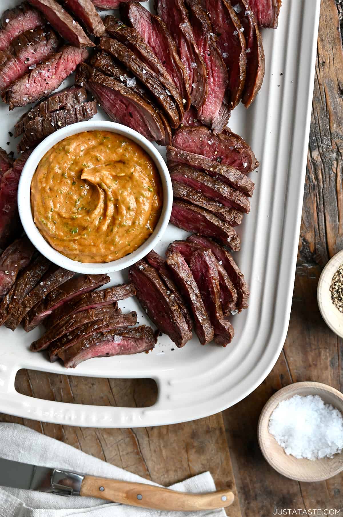 A top-down view of a small bowl containing cowboy butter on a serving platter surrounded by sliced grilled steak.