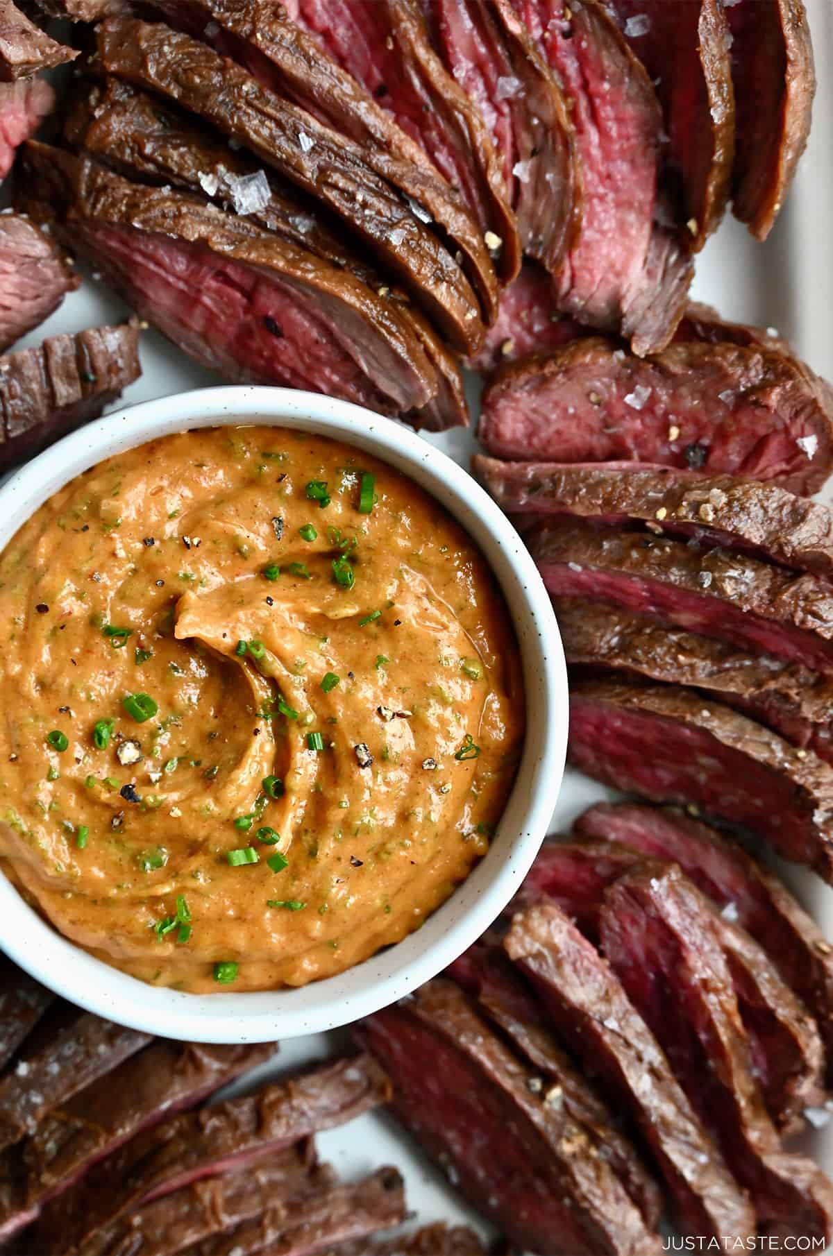 A close-up view of cowboy butter in a small white bowl on a plate surrounded by sliced flank steak.
