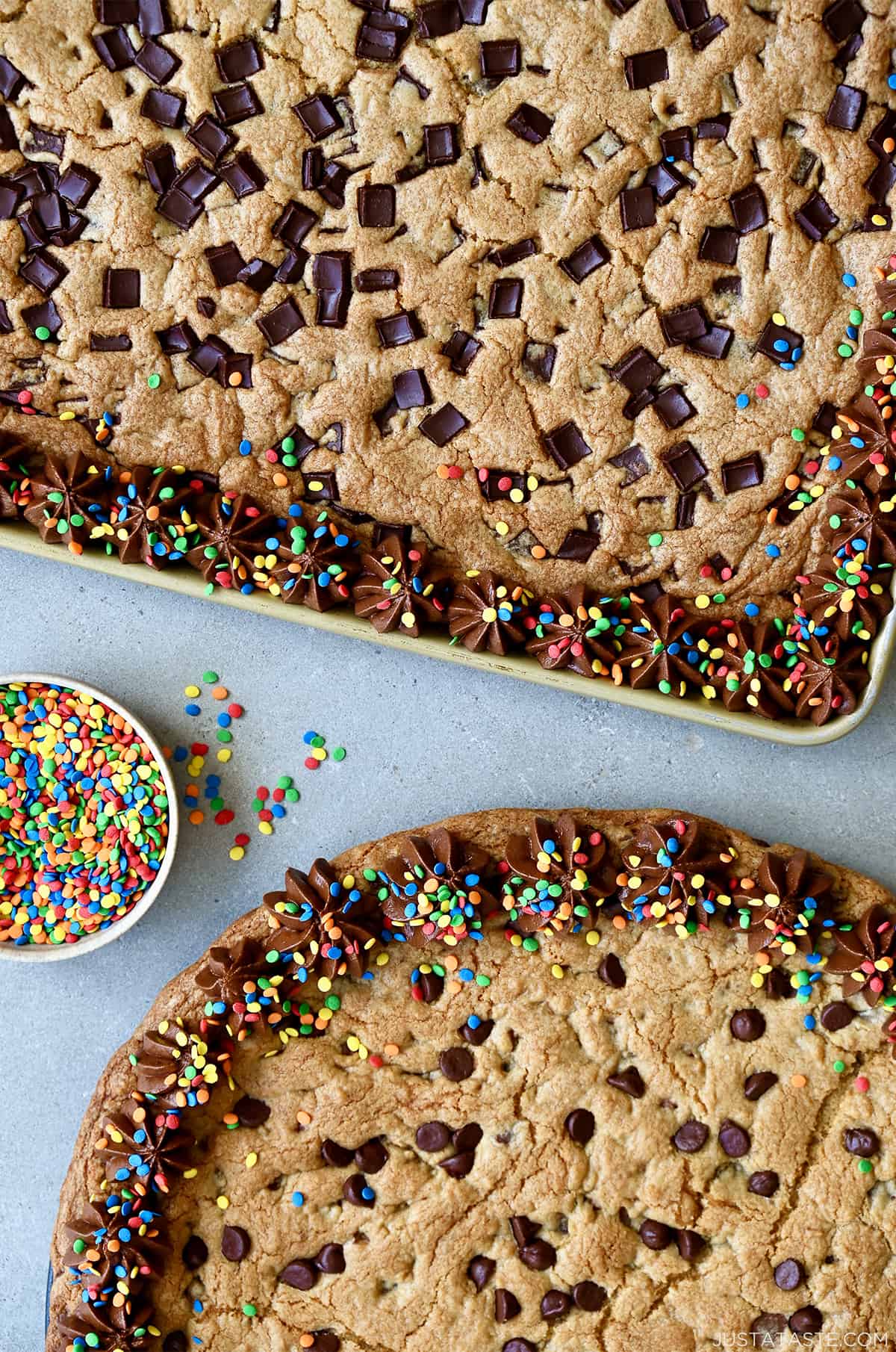 A top-down view of two cookie cakes studded with chocolate chips and chunks, plus chocolate buttercream frosting and rainbow sprinkles.