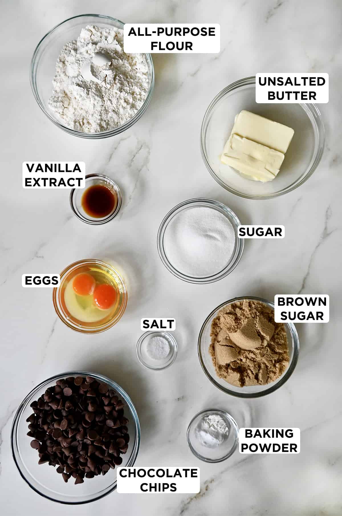 A top-down view of various sized bowls containing flour, butter, sugar, brown sugar, salt, eggs, vanilla extract, chocolate chips and baking powder.
