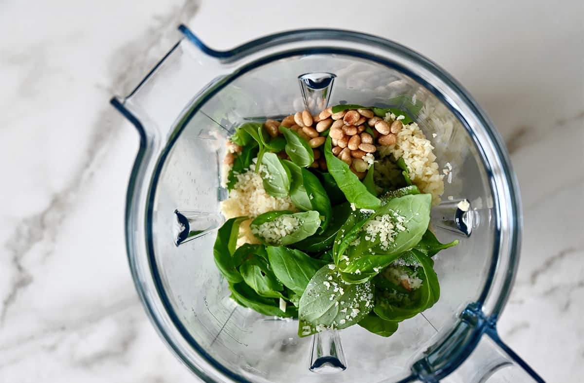 A top-down view of a blender container with fresh basil leaves, pine nuts, garlic and Parmesan cheese.